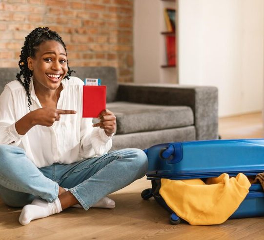 smiling-black-woman-pointing-passport-with-air-tickets-preparing-journey-sitting-near-packed-suitcase-home_116547-52886