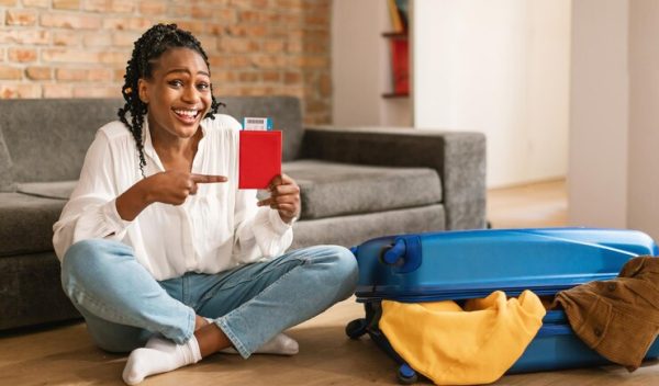 smiling-black-woman-pointing-passport-with-air-tickets-preparing-journey-sitting-near-packed-suitcase-home_116547-52886