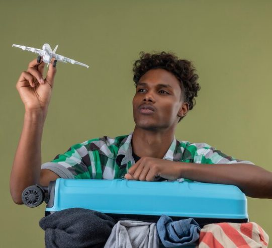 young-african-american-traveler-man-with-suitcase-holding-toy-air-plane-looking-it-with-serious-confident-expression-face-green-background_141793-30047