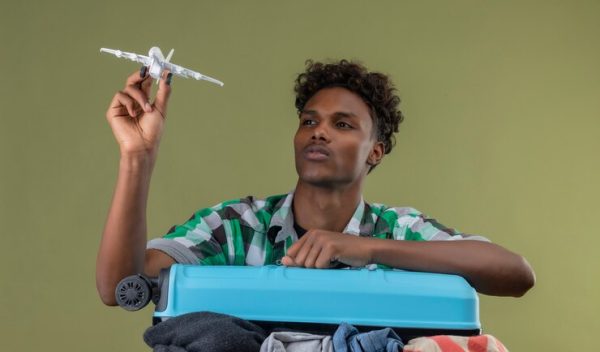 young-african-american-traveler-man-with-suitcase-holding-toy-air-plane-looking-it-with-serious-confident-expression-face-green-background_141793-30047